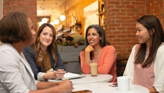 Fyra kvinnor samtalar runt ett cafébord med anteckningsbok